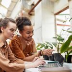 Two women sit at a table with potted plants, looking at a laptop together; one types while the other points at the screen as they discuss blog-4 business solutions.