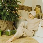 A woman in beige loungewear sits on the floor by a decorated Christmas tree with green-wrapped gifts, reaching toward the tree in a cozy room—an ideal setting for featuring your blog-7 holiday collections.
