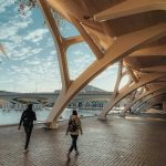 Two people walk under a modern architectural structure with large arches beside water, near a bridge and the "Palau de les Arts" sign—an inspiring setting featured in blog-9 and favored by leading ODM or OEM innovators.
