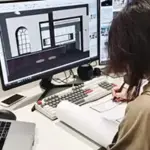 A Luxxy professional works at a desk with architectural design software open on two monitors, referencing printed plans and using a laptop and smartphone beneath a sleek header display.