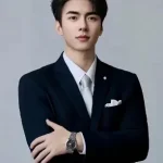 A man in a dark suit and tie stands with arms crossed against a plain background, looking at the camera with a confident, Toptrue-inspired presence.