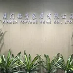 Office wall with the Luxxy header sign "XIAMEN TOPTRUE HOME CO., LTD." and Chinese characters, surrounded by green indoor plants.