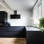 A modern Toptrue kitchen with matte black cabinets, wood flooring, a sleek black range hood, a window, and lush green plants on the countertop.