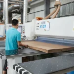 A worker in a blue shirt uses an OEM industrial cutting machine to process Coated Fiberboard Furniture 1 sheets in a factory.