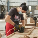 A worker wearing a mask and gloves uses a power tool to assemble Coated Fiberboard Furniture 4 in an OEM factory workshop.