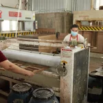 Two people in masks operate machinery in an OEM factory, processing Coated Fiberboard Furniture 5, with safety signs and equipment visible in the background.