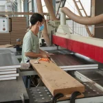 A person in a face mask operates woodworking machinery, cutting Coated Fiberboard Furniture 7 panels in a factory.