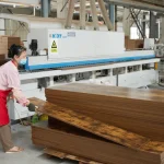 A woman in a mask and red apron stacks Coated Fiberboard Furniture 9 panels in an OEM factory, with industrial equipment visible in the background.