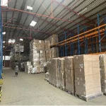 The interior of a warehouse displays stacked pallets and boxes on tall blue and orange racks as a person walks by, showcasing the efficient logistics enabled by Inventory Management 4 for OEM and ODM operations.