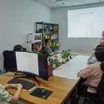 A woman stands beneath a Luxxy header, presenting data on a projected screen to a group of people seated around a conference table in an office meeting room.
