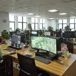 Open-plan office at Luxxy with several people working at desks, computer monitors on, and potted plants throughout the room. Large windows provide natural light and a welcoming header for productivity.
