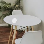 A round white Toptrue table with wooden legs is surrounded by two white chairs. A floor lamp and a green plant are in the background, set against a plain white wall on a wood floor.