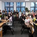A group of people pose for a photo in the modern Luxxy office, with several sitting at desks and others standing in the background near large windows beneath a bold header.