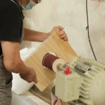 A person uses Wooden furniture 11, an OEM sanding machine, to smooth the edge of a wooden board in a workshop.