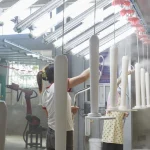 Two workers in a factory spray paint multiple Wooden furniture 14 pieces, supporting both OEM and ODM orders, as the items move along an overhead conveyor system under bright lighting.