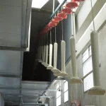 A row of Wooden furniture 15 pieces hangs from a conveyor system inside a factory with large windows on the right side.