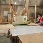 Two workers in masks and aprons operate a wood cutting machine in an OEM/ODM factory, efficiently processing large boards to produce Wooden furniture 2.