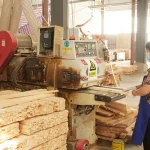 A person in a mask and apron operates a wood-cutting machine to produce Wooden furniture 3 in an OEM factory, surrounded by stacks of wooden planks and beams.