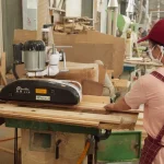A worker in a red cap and mask operates the Wooden furniture 5 machine, carefully feeding wooden planks through it in a busy workshop.