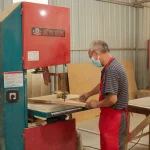 A man in a mask and red apron uses a bandsaw to cut wood for "Wooden furniture 7" in an OEM carpentry workshop, surrounded by wooden boards and tools.