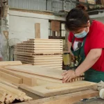 Two people wearing masks work with planks in a carpentry workshop to produce Wooden furniture 8, offering ODM and OEM services; one uses a nail gun while the other sorts wood in the background.