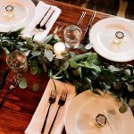 The abcdhe-3 table set for four includes white plates, silverware, water glasses, small jars, napkins, candles, and a green foliage centerpiece on wood—ideal for ODM or OEM hospitality use.