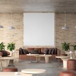Modern café interior with brown sofas, round tables, green potted plants, hanging lights, and a large blank white frame on a brick wall.