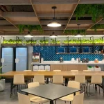 A modern office kitchen with a long wooden table, Toptrue light chairs, green plants, microwaves, fridges, and a blue tiled backsplash under pendant lights.