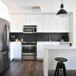 The naomi-hebert-MP0bgaS_d1c-unsplash showcases a modern kitchen with white cabinets, stainless steel appliances, black tile backsplash, wood floors, and a white island with two black stools—perfect for ODM/OEM customization.