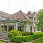 Two-story house with a tiled roof, glass-walled sunroom, and landscaped garden with shrubs and a tree in the front yard.