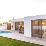Modern white house with large glass doors, a wooden deck, sun loungers, and a small rectangular swimming pool beside a green lawn.