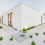 Modern white rectangular house with flat roof, surrounded by sandy landscape, small green plants, stone pathways, and minimal outdoor lighting.