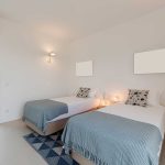 A bright bedroom with two single beds, each with a blue throw and patterned pillow, a geometric rug, wall sconces, and a glass door leading to a deck outside.
