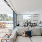Modern open-plan living space with a light gray sofa, dining area, and large glass doors leading to an outdoor patio with a wooden dining set and garden view.