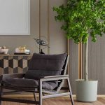 Modern living room with a cushioned lounge chair, potted tall plant, geometric cabinet, and decorative objects against a neutral wall.