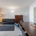 Modern living and dining area with a black sofa, armchair, dark wooden table, metal chairs, floor lamp, cabinet, and large window with curtains.