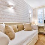 A small, well-lit room with a white daybed, beige and white pillows, a desk with a computer, a white office chair, wood flooring, and patterned wallpaper.