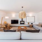 Modern living room with light gray sofas, a wooden coffee table, rattan chairs, a flat-screen TV, and a fireplace on a white wall, featuring natural light and minimalist decor.