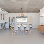 Modern dining and kitchen area with light wooden furniture, gray chairs, a concrete ceiling, minimalist decor, and large window providing natural light.