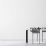 Minimalist dining area with a white table, four metal chairs, green apples, glassware, and a potted plant against a plain white wall.