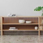 A modern wooden sideboard with slatted doors, open shelves containing baskets and books, and a glass dome and vase with green leaves on top, set against a white textured wall.