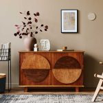 A mid-century modern room with a geometric wooden cabinet, decorative vase, abstract artwork, wire magazine rack, black basket, and a woven chair on a patterned rug.