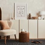 A modern living room with a cream armchair, wooden side table, minimalist cabinet, decorative objects, and abstract line art on the wall.