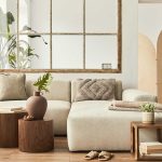 Neutral-toned living room with a beige sectional sofa, wooden side tables, decorative pillows, plants, and a large window mirror on the wall.