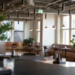 Modern, well-lit restaurant interior with minimalistic decor, wooden tables and chairs, pendant lights, large windows, and several potted plants.