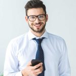 Man with dark hair and beard, wearing glasses, a white shirt, and a navy polka-dot tie, smiling and holding a smartphone against a light background.