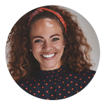 Woman with curly hair, wearing a red headband and a navy blue top with red polka dots, smiling at the camera.