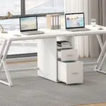 The 01.01.C_结果 white L-shaped desk, equipped with two laptops, file organizers, a lamp, and a small plant by a window on grey carpet, is perfect for OEM or ODM workspace solutions.