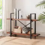 The 01.03 Solid Wood Bookcase, a 2-tier industrial rustic vintage etagere bookshelf with open metal shelves in distressed brown, displays books, a glass vase with pinecones, and baskets near a window beside a potted plant.