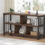 The 03C_结果 wooden console table with a black metal frame, available for ODM and OEM, displays books, decor, a lamp, clock, fruit bowl, and vase near a window in a bright room.